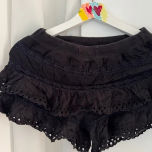 Svarta volangshorts med broderade detaljer - Supersöta svarta shorts med flera lager volanger och broderade spetsdetaljer. De har en bred resår i midjan och är perfekta för en trendig och avslappnad look.😍
