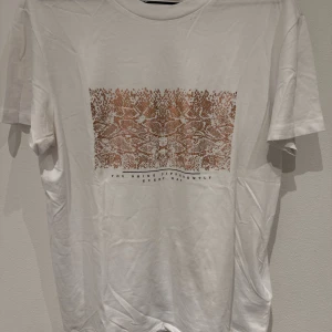 Vit t-shirt med ormskinnsmönster - Vit t-shirt med ett stort brunt ormskinnsmönster på bröstet och texten 'You shine differently every day' under motivet. Klassisk passform och rund halsringning. Perfekt för dig som vill sticka ut lite extra.