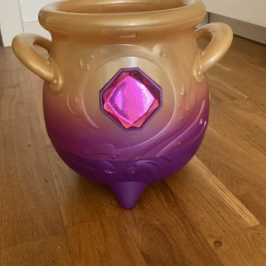 Magic Mixies med lila och beige färg kommer med docka och en stav. - Säljer en stor Magic Mixixes i beige och lila med glittrig rosa ädelsten på framsidan. Kitteln har två handtag och dekorativa stjärnor samt ett reglage för olika spellägen. Perfekt för magiska lekar och rollspel.