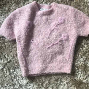 Ljusrosa fluffig tröja från Zara - Superfin ljusrosa stickad tröja från Zara med korta ärmar och fluffig textur. Tröjan har dekorativa detaljer framtill som ger en unik look. Perfekt för dig som gillar mjuka och mysiga plagg!