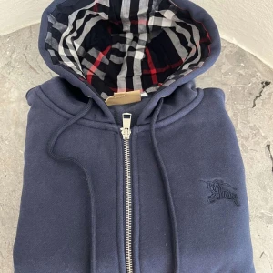 Blå huvtröja från Burberry - Säljer en snygg blå huvtröja från Burberry med dragkedja och broderad logga på bröstet. Insidan av huvan har ett klassiskt rutigt mönster i svart, vitt och rött. Perfekt för en stilren look. Pris går även att diskutera 