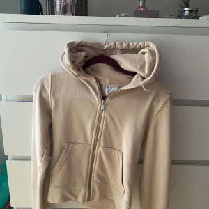Beige hoodie med dragkedja från Divided - Säljer en beige hoodie från Divided i storlek XS. Tröjan har dragkedja framtill, huva och två fickor. Perfekt för en avslappnad stil och enkel att matcha med det mesta. Aldrig använd