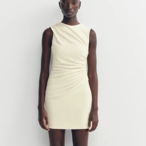 Krämvit kort klänning med drapering - Aldrig använd!!Snygg krämvit kort klänning med asymmetrisk drapering i midjan och axelpartiet. Ärmlös modell med rund halsringning och figurnära passform. Perfekt för dig som gillar stilrena och moderna plagg.