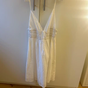 Vit sommarklänning med smock och knytband - Säljer en söt vit klänning med smala axelband, v-ringning och smock i midjan. Klänningen har knytband framtill och är tillverkad i ett lätt material, perfekt för varma dagar.
