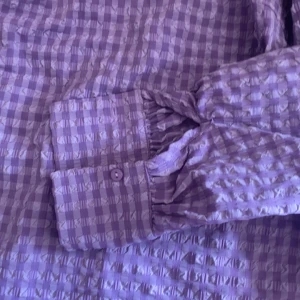 Lila rutig skjorta med struktur - Säljer en lila skjorta med rutigt mönster och strukturerad yta. Skjortan har långa ärmar med manschett och knappar samt en klassisk krage. Perfekt för dig som vill ha något unikt och stilrent i garderoben. (Problem med bakåtknappen)