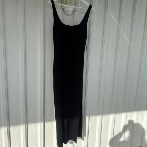 Svart långklänning med smala axelband - En stilren svart långklänning med smala axelband och rundad urringning. Klänningen har en rak passform och är ärmlös, perfekt för en enkel och elegant look.