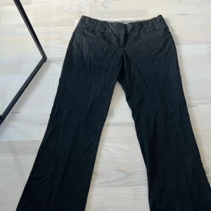 Svarta kostymbyxor från Monsoon - Klassiska svarta kostymbyxor från Monsoon med raka ben och diskreta knappar vid midjan. Byxorna har fickor bak och är perfekta för en stilren look. Passar dig som gillar enkel och tidlös design.