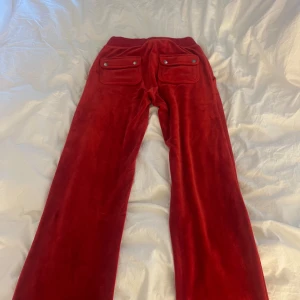 Röda Juicy Couture byxor  - Säljer ett par röda velourbyxor med bred resår i midjan och stora fickor med knappar bak. Byxorna har en rak och lite loose passform, perfekta för en avslappnad stil.