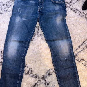 Blå jeansbyxor från Dsquared2 - Säljer ett par klassiska blå jeans från Dsquared2 med slitningar och ljusa partier. Byxorna har fem fickor, knappgylf och en röd Dsquared2-tag vid gylfen. Perfekta för dig som gillar en avslappnad och trendig look. Mycket fräscha och inga defekter, använts 4 gånger 