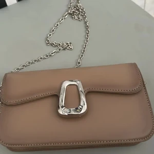 Beige axelväska från Rizzo med silverkedja - Säljer en stilren beige axelväska från Rizzo med silverfärgad kedja och dekorativt spänne framtill. Väskan har magnetlås och ett innerfack med dragkedja. Perfekt för dig som gillar klassisk och enkel design. Priset kan diskuteras om de går smidigt till. 