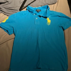 Blå piké från Polo Ralph Lauren str 164 cm - Säljer en blå pikétröja från Polo Ralph Lauren med stor gul logga på bröstet och siffran 3 på ärmen. Klassisk krage och knappar framtill. Perfekt för en sportig och stilren look.