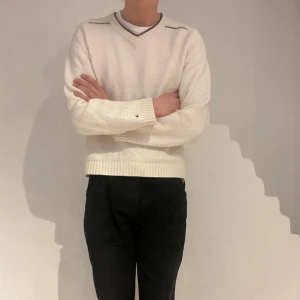 Vit stickad tröja från Tommy Hilfiger M/XL - Vit stickad tröja från Tommy Hilfiger med v-ringad hals och diskret logga på armen. Lite croppad passform med långa ärmar. Tröjan har ribbade muddar och brun detaljsöm längs axlarna. Gjord i bomull och lammull!     Herrstorlek M/L - Damstorlek XL