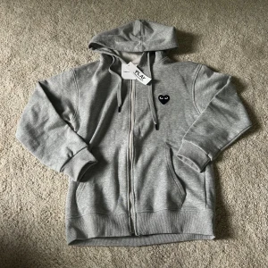 Grå Comme des Garçons Zip Hoodie - Ny och endast testad en gång, men passar inte. Enkel och stilren design som passar perfekt för en casual look.  Skick: Ny, testad en gång Storlek: M Färg: Grå  Hör av dig om du vill ha fler bilder eller info!