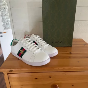 Gucci Ace vita sneakers med bi-detalj - Gucci Ace sneakers i vitt skinn med klassiska gröna och röda ränder på sidan och broderad guldfärgad bi. Baksidan har metallicröd och metallicgrön häl med Gucci-logga. Platt vit sula med unikt mönster undertill. Snygga och ikoniska sneakers för dig som gillar lyxig streetstyle.