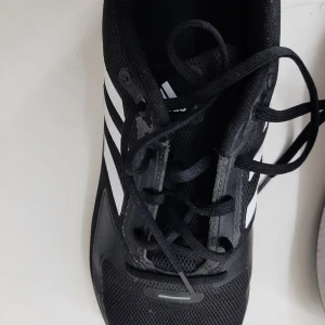 Svarta Adidas sneakers med vita ränder - Svarta Adidas sneakers med klassiska vita ränder på sidorna och vit sula. Ovandelen är i mesh för bra andningsförmåga och skon har snörning. Perfekta för träning eller en sportig look. Snygg och stilren design som funkar till det mesta.