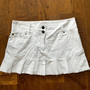 Vit kortkjol med volangkant - Snygg vit kortkjol med volangkant och råa kanter längst ner. Kjolen har två knappar framtill, fickor både fram och bak samt coola sömdetaljer. Perfekt för dig som vill ha en fräsch och stilren look med lite edge.