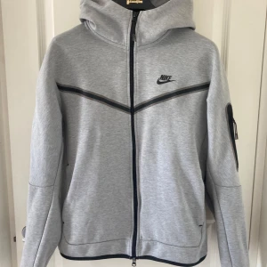 Grå Nike tecch flecce - Mycket bra skick ! Snygg grå tecch fleece från Nike med svart dragkedja och svarta detaljer över bröstet. Jackan har huva, lång ärm och en extra ficka med dragkedja på ena ärmen. Perfekt för en sportig och avslappnad stil.