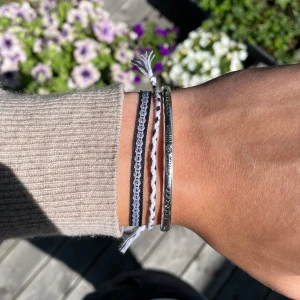 Svartvitt armband i silver och flätade armband - Snyggt armband med flera band i svart, vitt och silver. Kombination av flätat tyg och tunna silverfärgade metallband med graverade detaljer. Perfekt accessoar för en cool och avslappnad stil. Justerbar knytning med tofsar för personlig passform.