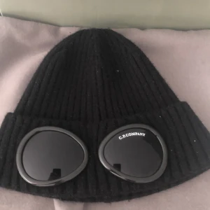 Svart C.P. Company Goggle Beanie - Svart ribbstickad mössa från C.P. Company med ikoniska goggle-linser framtill. Mössan är tillverkad i mjuk ull och har en uppvikt kant. Perfekt accessoar för dig som vill sticka ut med streetwear-vibe.