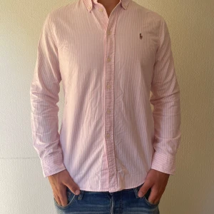 Rosa randig skjorta från Ralph Lauren - Säljer en klassisk rosa och vit randig skjorta från Ralph Lauren med broderad logga på bröstet. Skjortan har lång ärm, knappar framtill och en snygg krage. Perfekt för dig som gillar stilrena och fräscha plagg i bomull.