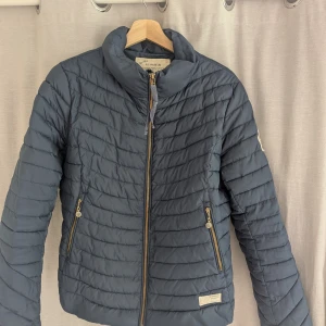 Mörkblå dunjacka från Odd Molly  - Mörkblå pufferjacka Odd Molly Downfall Jacket med quiltad design och guldfärgade dragkedjor. Jackan har hög krage, två fickor med dragkedja och en snygg patch på ena ärmen. Perfekt för kyliga dagar och riktigt skön att bära.