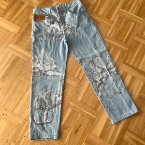Ljusblå jeans med änglamotiv från DV - Unika ljusblå jeans från Don Vito med detaljerade svarta änglamotiv tryckta över hela byxan. Klassisk femficksmodell med broderad DV-logga på bakfickan och läderpatch framtill. Raka ben och normal passform, perfekt för dig som vill sticka ut.