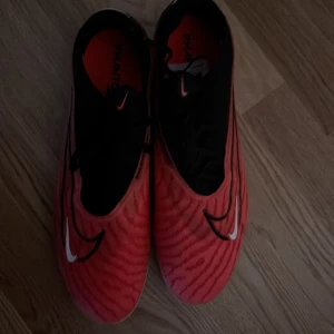 Nike röda fotbollsskor Phantom GT - Säljer ett par Nike Phantom GT fotbollsskor i rött och svart med vita detaljer. Skorna har snörning, mönstrad ovandel och dobbar för gräsplan. Snygg design med svarta och vita Nike-loggor och coola vågmönster på ovandelen.