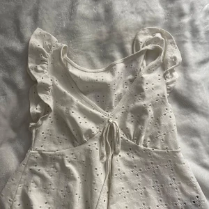 Vit somrig blus med volangärmar - Superfin vit blus med broderade hålmönster, volangärmar och v-ringning med söt knytning framtill. Lätt och luftig, perfekt för varma dagar. Blusen har en feminin och romantisk stil med fina detaljer.