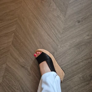 Svarta kilklack-sandaler från Högl - Snygga svarta sandaler från Högl med bred rem i skinn och öppen tå. Skorna har en hög korkliknande platåklack och justerbar rem runt hälen. Perfekta för dig som vill ha både stil och höjd på sommaren.