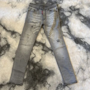 Grå skinny jeans från Dsquared2 - Grå skinny jeans från Dsquared2 med coolt slitna detaljer och färgstänk. Klassisk femficksmodell med Dsquared2-logga på röd etikett och stor röd patch bak i midjan. Jeansen har smal passform och är tillverkade i stretchig denim för extra komfort.