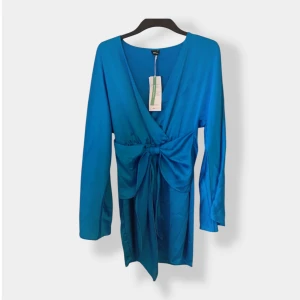 Blå omlottklänning från Gina Tricot - Supersnygg blå kort omlottklänning från Gina Tricot med lång ärm och knyt i midjan. Klänningen har v-ringning och är gjord i ett glansigt polyestertyg som ger en lyxig känsla. Perfekt för dig som vill sticka ut med färg och stil.