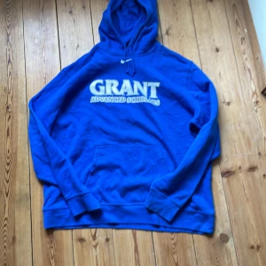 Blå Nike hoodie Grant XXL - Snygg blå hoodie från Nike med stor vit text 'GRANT ADVANCED SCHOLARS' på bröstet. Klassisk huva med snörning och magficka framtill. Perfekt för dig som gillar oversized streetstyle och vill sticka ut med en färgstark look.