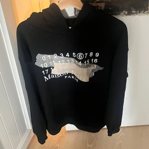 Maison Margiela hoodie - Svart hoodie från Maison Margiela (Passar L men är M) med ikoniskt sifferprint och tejpdetalj på bröstet. Tröjan har huva och är tillverkad i mjuk bomull. Min leverantör säger att den är äkta, men jag är misstänksam därför det låga priset skriv vid ytterligare funderingar🙌