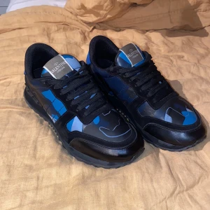 Valentino Garavani Rockrunner sneakers - Snygga Valentino Garavani Rockrunner sneakers i svart och blått med coolt kamouflagemönster. Skorna har svarta snören, rund tå och detaljer i skinn och mocka. Ikoniska nitar baktill på sulan ger en edgy vibe. Perfekta för dig som vill sticka ut med stil. Riktigt fint skick men man bedömer själv efter bilderna!(INGET OG MEDFÖLJER) självklart äkta! Hör av er vid minsta lilla 