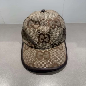 Gucci beige keps med GG-mönster - Snygg keps från Gucci i beige och brunt med det klassiska GG-monogrammet över hela kepsen. Kepsen har böjd skärm, justerbar rem baktill i brunt och är tillverkad i bomullscanvas med detaljer i skinn. Perfekt för dig som vill sticka ut med en lyxig accessoar.