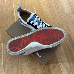 Christian Louboutin sneakers med nitar - Snygga sneakers från Christian Louboutin med ikonisk röd sula, vita och svarta paneler, blå detaljer och coola silverfärgade nitar på tån. Skorna har snörning och är tillverkade i skinn med inslag av mocka. Perfekta för dig som vill sticka ut med din stil.