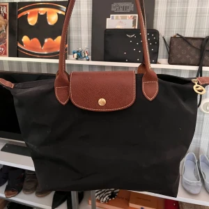 Svart Longchamp axelväska med skinn - Klassisk svart axelväska från Longchamp med bruna skinnhandtag och lock. Väskan har guldfärgad knapp och dragkedja, samt rymlig insida. Perfekt storlek för att få plats med allt du behöver och enkel att bära över axeln.