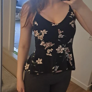 Svart blommig linnetopp med volang - Svart linnetopp med blommor. Toppen har smala axelband, v-ringning och en lätt utsvängd volang nertill. Ställ gärna frågor! 💕