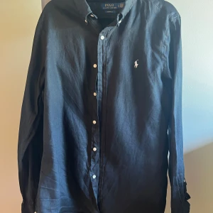 Svart skjorta från Polo Ralph Lauren - Klassisk svart linne skjorta från Polo Ralph Lauren i custom fit. Skjortan har långa ärmar, knappar framtill och den ikoniska broderade loggan på bröstet. Tillverkad i linne, perfekt för en stilren look.