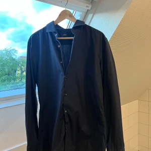 Hugo boss mörkblå skjorta - Mörkblå skjorta från Hugo Boss i slim fit. Sitter riktigt snyggt och passar till både fest, skola eller om du bara vill klä upp dig lite. Easy iron också, så den är enkel att stryka. Använd ett fåtal gånger och är i jättebra skick. Storlek 41, vilket motsvarar ungefär M/L. Nypris låg på runt 1200–1300 kr. Säljer bara för att den inte passar mig längre. Pris kan diskuteras!