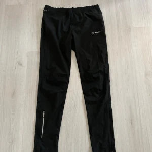 Svarta träningsbyxor från SOC - Svarta träningsbyxor från SOC med resår och snörning i midjan. Lätta och vindtäta med TCS Wind-teknologi, perfekta för löpning eller utomhusträning. Reflekterande detalj vid benet för extra synlighet. Snygg och sportig look.