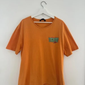 Orange t-shirt med Paradise-tryck - Snygg orange t-shirt med coolt tryck fram och bak. På bröstet står det 'Paradise' i grön graffiti-stil och på ryggen finns ett stort motiv med öken, kaktusar, dödskalle och hästsko. T-shirten har rund hals och korta ärmar, perfekt för dig som gillar unika prints.