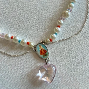 Pärlhalsband med hjärthänge och vintagebild - Unikt halsband med vita pärlor, små blå och röda pärldetaljer samt ljusrosa pärlor. I mitten sitter en oval berlock med vintagebild av en kvinna i röd klänning och ett genomskinligt hjärthänge i ljusrosa. Silverfärgad kedja ger extra lager.