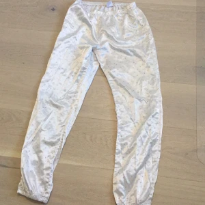 Vita satinbyxor med hjärtan KappAhl - Snygga vita byxor från KappAhl i glansigt satinmaterial med små rosa hjärtan som mönster. Byxorna har resår i midjan och vid bensluten för en bekväm passform. Perfekta för dig som gillar mjuka och sköna byxor med söt detalj.