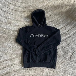 Svart Calvin Klein hoodie med logga - Svart hoodie från Calvin Klein med stor vit logga framtill. Klassisk modell med huva och känguruficka, ribbade muddar och lång ärm. Perfekt för en chill och stilren look. Materialet är mjuk bomullsmix som känns skönt mot huden.