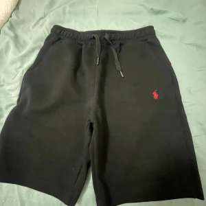 Svarta shorts från Polo Ralph Lauren - Svarta shorts från Polo Ralph Lauren med röd broderad logga på vänster ben. Mjuk bomullskänsla, elastisk midja med snörning och två sidofickor. Perfekta för chill dagar eller när du vill ha en clean och klassisk look.