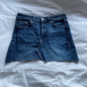 Jeanskjol med spetsdetaljer - Kort jeanskjol i klassisk blå denim med rå kant nertill och vita spetsdetaljer på bakfickorna. Har sytt om ett bar jeans till kjol. Har en nagellims fläck på ena bakfickan som ej har gått bort.