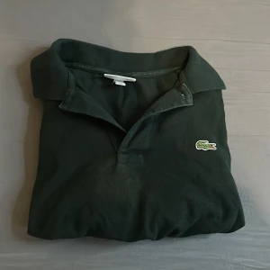 Mörkgrön pikétröja från Lacoste L - Snygg mörkgrön pikétröja från Lacoste i klassisk modell med krage och korta ärmar. Ikoniska krokodilloggan broderad på bröstet. Tillverkad i bomull och passar perfekt för en clean och sportig stil. Hör av er vid minsta lilla fråga. 