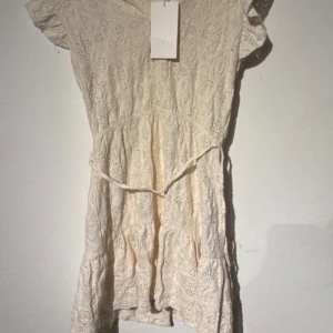 Vit spetsklänning från Zara S - Supersöt vit kortklänning från Zara i spets med knappar framtill och knytband i midjan. Klänningen har korta ärmar och volang nertill, perfekt för en fräsch och somrig look. Materialet är bomull och ger en luftig känsla.