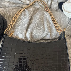 Svart axelväska med kedja - Snygg svart axelväska i croco-mönstrat skinnimitation med bred guldfärgad kedja som axelrem. Väskan har dragkedja upptill och en stilren, rektangulär form. Perfekt för dig som vill ha en trendig accessoar med lyxig känsla.